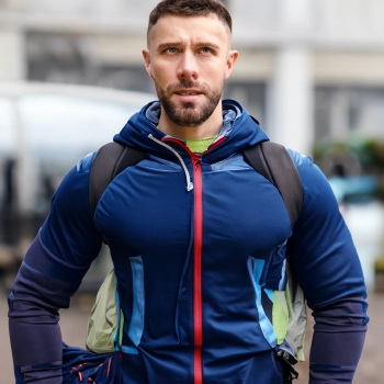 A muscular man wearing blue jacket standing alone with a back pack