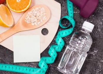 A table with a measuring tape, bottle of water, oatmeal, fruits, and a dumbbell