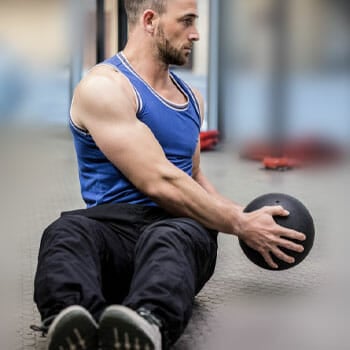 man in a tank top working out his core with a medicine ball