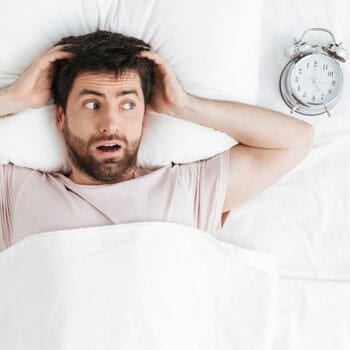 man laying down in bed in panic with his alarm clock beside