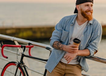 A man relaxing near a bridge beside his bike while holding a coffee