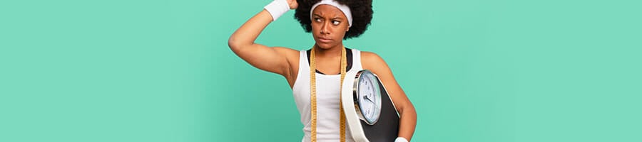 black woman in gym clothes with measuring tape on her neck and holding a scale thinking