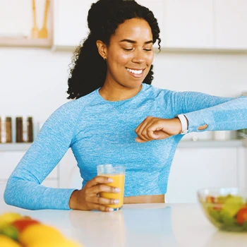 A woman drinking a fruit drink while looking at her smart watch in the kitchen