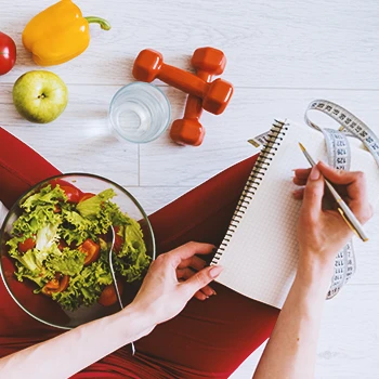 A woman writing down notes with a salad on her lap and workout equipment in front