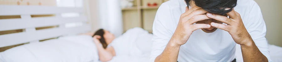 A stressed man while sitting in bed with a woman in the background